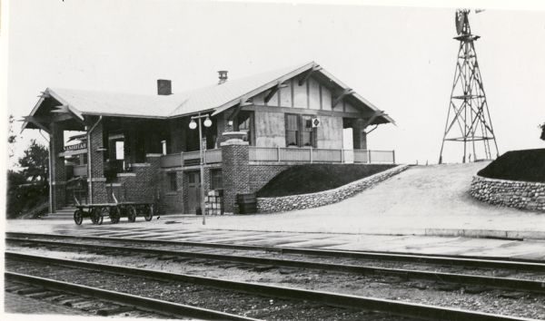 Grayscale long shot of the Nashotah Railway Depot in the distance, sitting on sloping terrain. Railway tracks stretch from left to right foreground. The depot is on the left background. The building's side that faces the railroad tracks features a grand staircase and a portico with two supportive columns, among others. Two empty baggage wagons stand between the tracks and the depot on the left. On the right of the depot is a steep driveway leading up to a windmill.
