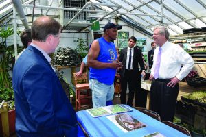 Wide shot of Will Allen in a sleeveless blue Growing Power t-shirt talking to a visitor from the U.S. Department of Agriculture on the right inside a greenhouse. Allen stands in the center of the image. Other people in suits surround Allen and his visitor. Designs in a binder are placed on a blue table below Allen, and plants are visible in the background.