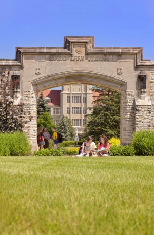 Long shot of two students in T-shirts sitting on a green lawn in the distance. Behind them is a grand arched stone gateway that is inscribed "Mount Mary College" on the top front. Three students gather below it. A university building is visible in the far background. A green lawn spans in the foreground. Above is a clear blue sky.