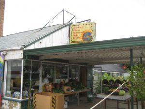Photograph of a covered porch with tables full of various vegetables. The porch is set on a single-story building that features glass window walls and an entrance. Atop the porch's roof is a sign that reads "Growing Power Community Food Center" with a web address below.