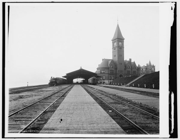 Grayscale long shot of the Chicago and North Western rail depot. The center portion of this image displays long railroad tracks headed toward a shelter at the center of the photograph. The train depot appears in the background with a tall clock tower on the right.