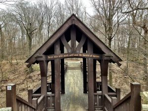Photograph of the wooden roofed Grant Park entrance with handrailings along its sides towards a wooden pedestrian bridge. Text inscribed on a truss beam below the roof reads, "Enter this wild wood and view the haunts of nature." Leafless trees appear here and there around the bridge and in the background.