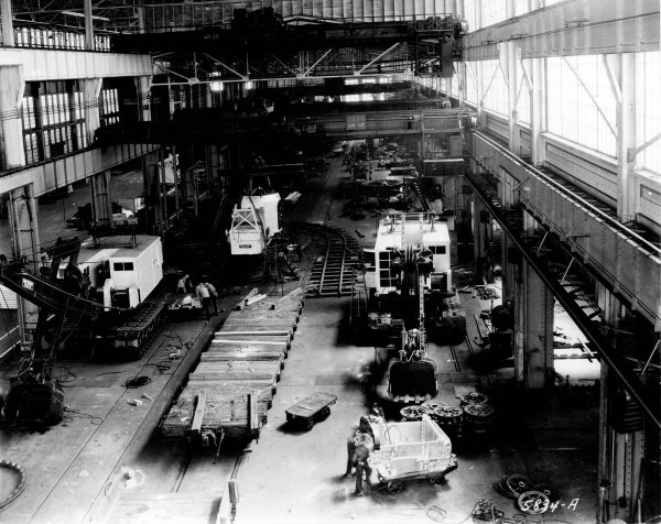 High-angle shot of the interior of the Bucyrus-Erie plant in grayscale. Rays of sunshine from large windows on the right light up heavy equipment, gears, and other machines lying on the floor. Some people work down there. Steel structures are visible in the background.