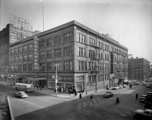 Long shot of the corner view of T.A. Chapman's department store by a street corner in grayscale. The facade is on the left. Another side of the building is on the right. The front section consists of a four-story structure featuring the main entrance, two marquee awnings, a vertical store sign, and several display windows on the ground floor. The store name is also painted on the exterior wall of an adjacent building's top side on the left. People walk the sidewalk around the department store. A bus and cars traverse the street. Some vehicles are parked next to the store.