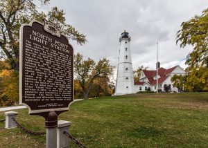 Long shot of the North Point Lighthouse and museum. The white-colored tower soars in the background. Its multiple windows, an observation deck, and a lantern room are visible in the distance. Next to it is the multiple-story museum with red roofs and white exterior walls. Several trees begin to turn with fall colors appearing dominantly on the left background. A large green yard is situated in front of the buildings and stretches toward the foreground. The North Point Light Station's historical marker stands on the lawn in the left foreground, facing slightly to the right. Above is a cloudy sky.