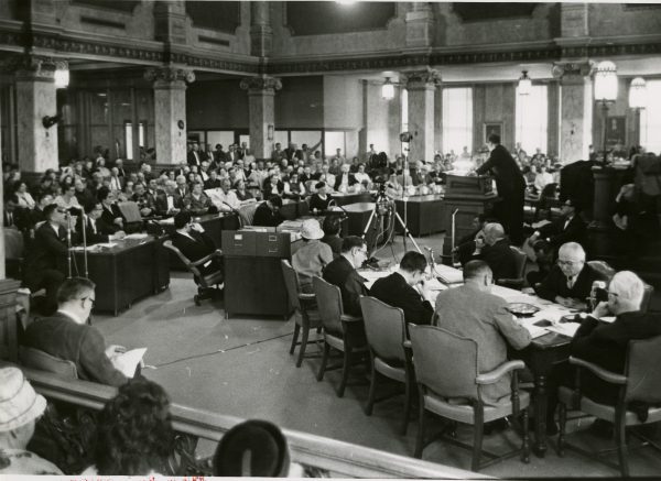 A man speaks at a podium to a crowd gathered for a hearing in regards to the Midtown public housing plant in the Milwaukee Common Council chambers in 1967.