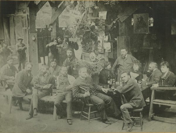 Sepia-colored group photo of the Milwaukee's panoramic painters sitting around a long table with bottles of beer on it. Richard Lorenz is seated farthest left behind the table. Some people stand a couple of feet behind the table.