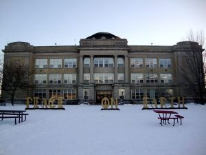 Long shot of Shorewood High School's three-story building behind three standing signs that read "Peace on Earth." The signs are set on the snowy ground. Two picnic tables are visible on the ground.
