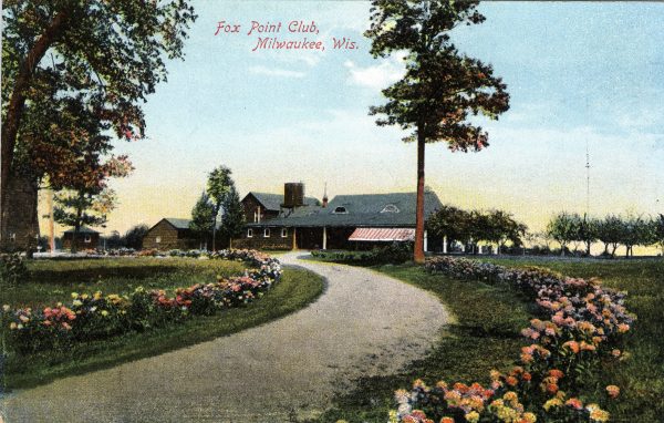 Postcard illustrating a panoramic view of the Fox Point Club. It features a building complex set among a large and well-maintained park. A pathway to the main entrance is shown in the center of the image. Colorful landscaping flowers grow along the path.