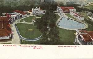 A painted postcard showcases a bird's eye view of the Wonderland amusement park. Its buildings with red-colored roofs surround an extensive green lawn. Lush green trees grow on the lawn and in the area behind the park. A water slide and a pool are visible on the postcard's right portion. Text at the bottom reads "Panoramic View, "Wonderland on the River, Milwaukee."