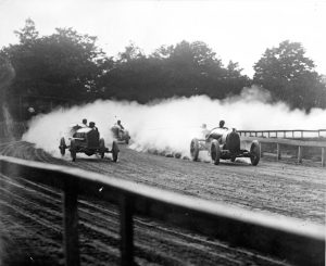 Wide shot of at least five antique cars speeding on dirt-surfaced track. This image's upper part shows a cloud of dust thrown into the air behind the automobiles driving through the corner of the circuit. The bottom portion of this grayscale picture displays a blurry image of the circuit fence.