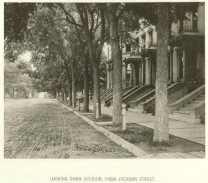 Sepia-colored image of Juneau Avenue stretching down on the left next to a line of trees growing on the road verge. Several seemingly identical dwellings with stone staircases and pillared entrances are visible on the far right. Text under the photo reads "Looking Down Division, From Jackson Street."