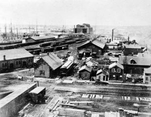 Grayscale elevated view of the first Milwaukee railroad depot and its surrounding area. The trains and railroad depot are in the background. Boats appear in the distant left. Buildings of different sizes are visible here and there around the depot area. A road stretches from left to right in the foreground.