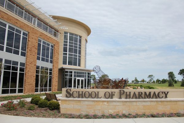 Long shot of the School of Pharmacy's facade against the cloudy blue sky. The modern design features exterior walls made of windows surrounded by brick. The school's monument sign stands on a stone base that appears from the center to the right portion of this image. A wire scuplture of a vintage pharmacist's mortar and pestle sits behind the monument sign. A group of landscaping plants surrounds the monument sign.