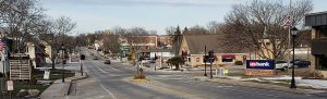 A panoramic view of Elm Grove's downtown features commercial buildings on either side of a long road in the middle of the image. Appearing in the foreground on the right is a U.S. Bank monument sign and the Plank Road Plaza standing signage on the left.