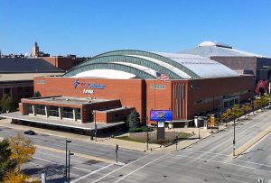 An elevated view of the UW-Milwaukee Panther Arena sitting on a street corner. The image showcases the building's sizeable arched roof and red bricks exterior walls. The old name sign that reads "U.S. Cellular Arena" is still attached to the top front of the structure. An intersection is in the image's foreground. Other buildings in the vicinity are visible.