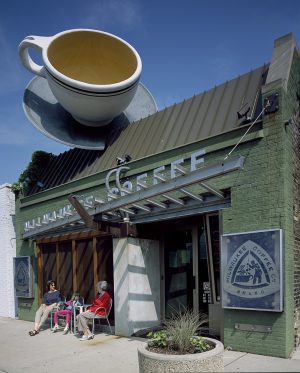 The façade of a coffee shop in Washington Heights with a large and tilted coffee cup-shaped ornament on its steep roof. The building faces slightly to the left against the blue sky. The one-story building's exterior wall is composed of bricks painted green. A tilted-designed name sign that reads "Milwaukee Coffee" is attached above the front windows and entrance. Two women and a young girl sit next to the windows. Two square store signs are set on the farthest left and right of the facade. A potted plant appears in the foreground.
