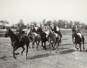 Grayscale full shot of a group of people in equestrian attire riding horses towards the left on an open ground at the Milwaukee Hunt Club. A golf course and tall trees are visible in the far background.