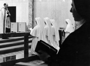 Grayscale wide shot of four nuns in white dress kneeling while facing left at the center background. A silhouette of another nun holding a bible appears dominantly on the right to the center front. A man in priestly garb stands at the left up a short flight of stairs.