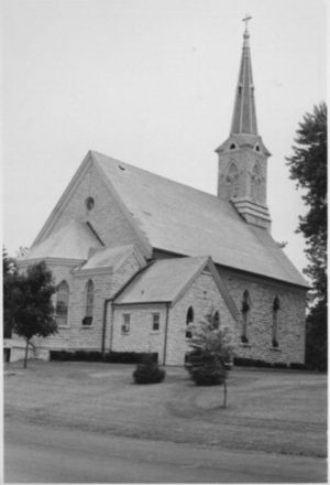 Sepia-colored long shot of Trinity Lutheran Evangelical Church exterior. The single-story building has a tower at the back with a cross atop. Three small trees grow on the lawn in the foreground.