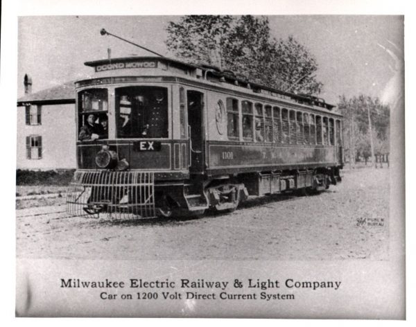 Grayscale wide shot of an interurban car with "Oconomowoc" sign on the top front part of its body. Some passengers are seated by the front window. A conductor with eight buttons on his jacket stands at the front. The car passes a building and trees in the background. The bottom of this photograph reads "Milwaukee Electric Railway & Light Company, Car on 1200 Volt Direct Current System."