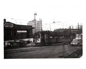 Sepia-colored long shot of an interurban streetcar running through a street from right toward left. A building with signage that reads "North Shore Line" sits by the street on the left. Two vintage cars appear on the road. Tall buildings are visible in the far background.