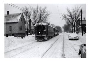 Long shot of the front view of an interurban streetcar traversing a snowy Milwaukee neighborhood. A long track stretches down next to the streetcar. Someone shovels the snow on the sidewalk on the left. Houses appear on the left side of the street. Snow covers the cars parked on the right.