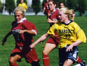 Full shot of two University of Wisconsin's women's soccer team players in red jerseys and their opponent from Marquette University in a yellow shirt running to the left. Kate Gordon of Marquette University is on the image's right. Her opponents are on the left. Other players and green trees are visible in the background. They play on a green field in daylight.