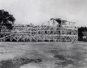 Grayscale wide shot of a massive roller coaster structure with some passengers riding on it. Two people stand below on the right, gazing up at the attraction.