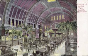 A painted postcard of the Schlitz Palm Garden's interior with an array of dining tables, palms and other plants scattered in the hall. The vaulted ceiling is colored in shades of purple. Inscribed in red ink on the top right of the postcard is "Interior Schlitz Palm Garden, Milwaukee."