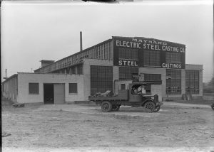 Long shot of Maynard Electric Steel Casting exterior in grayscale. The multiple-story structure has large signage on top and rows of industrial windows. On the left, adjacent to the building, is a single-story structure with a half-opened double door. A pick-up truck traverses the dirt road in front of the building.
