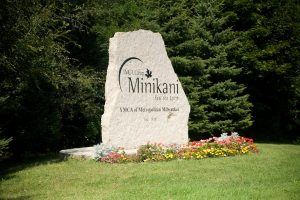 The YMCA Camp Minikani monument sign stands on a green lawn. Colorful landscaping flowers are set in front of the white-colored stone sign. Lush green bushes appear in the background.