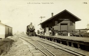 Long shot of the Dousman station of the Chicago and North Western Railway in sepia tone. It shows a crisscrossing railroad track line on the left and center of this image. To the right is the station's building, which features an open gabled roof and a chimney. Some people stand on the platform while a train approaches.