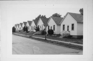 Grayscale long shot of the Carrolville Homes residential area showcasing several identical one-story dwellings lining from the left background to the right front. Each house features an entrance flanked by rectangular windows and a front stair connecting the door with the lawn. Some homes have trees in their front yards. A street, road verge, and sidewalk stretch from left to right foreground next to the residential places. A street sign that reads "No Outlet" stands on the road verge at the image's center.