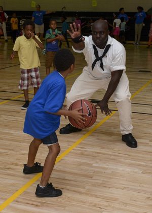 Image of a serviceman in his dress white uniform playing basketball with three children in an indoor basketball court. A child in the blue shirt holds the ball while facing the man. More children appear in the background along with other adults.