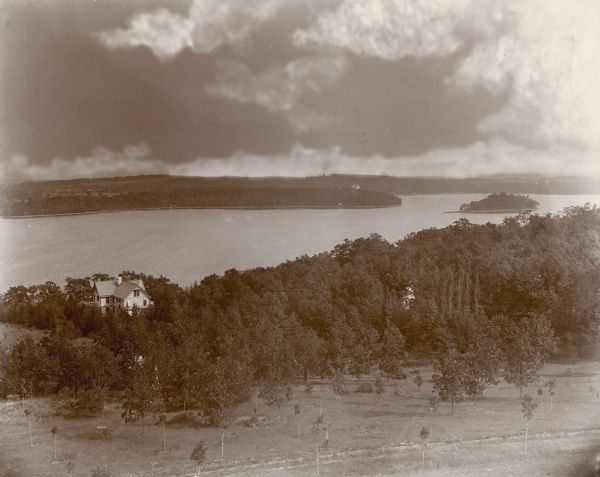 Panoramic view of Pine Lake and the centerpiece of the village of Chenequa in sepia tone. George Brumder's summer house appears in the distance among tall trees next to the large body of water.