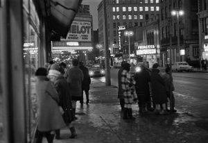 Back view of a group of people in warm clothes on a sidewalk in the foreground. Shining on the left background are lights from the underside of a building's marquee. Vehicles and street lamps glow on a roadway in the right background. Among the buildings lining the street's right side is the Princess Theater marked by its bright marquee.