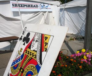Long shot of a banner reads "Sheepshead" hanging on the outside of a white-colored tent in the background. A gigantic printed art depicting the Queen of Clubs card appears in the image's foreground.