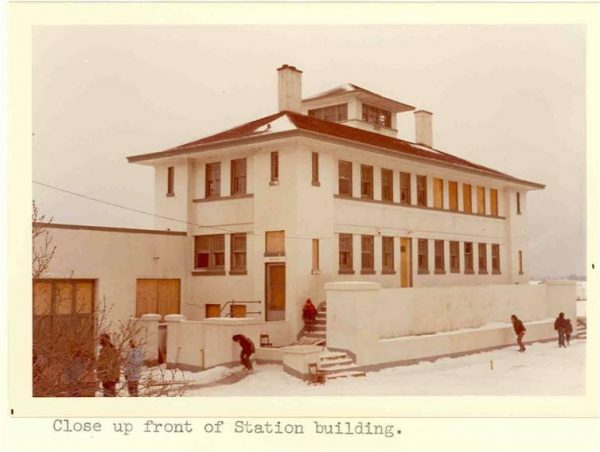 Wide shot of the facade of the McKinley Park Coast Guard Station. The two-story building has regularly spaced windows, light-colored walls, and a cupola on top. The station is surrounded by snow on the ground and on some parts of the roof. Some people appear outside the building, and one climbs the stairs to the station's entrance.