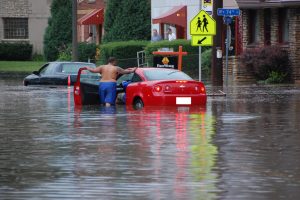 Long shot of a man in blue shorts opening the driver seat of a red car with a driver inside on a severely flooded street in Wauwatosa. The water is as high as his knees. Standing on their right are several street signs. In front of the red car is a black car partially submerged in floodwater. Standing on their right are several street signs. In the background, some people stand under red awnings installed on the dwellings' exterior. Green lawns, well-trimmed shrubberies and trees are visible next to the houses.