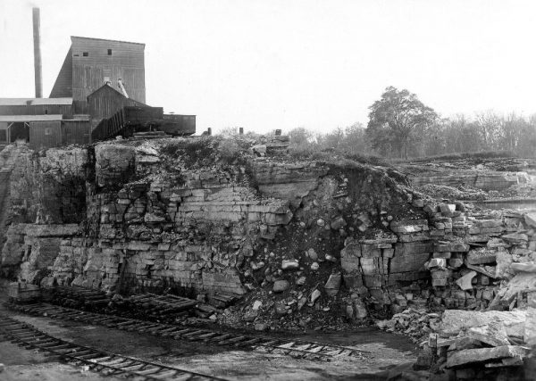 A grayscale photograph highlights a portion of the Lake Shore stone quarry. It shows a cross-section of rock and limestone. A wooden building and a smokestack appear in the upper left. Double-track railways are located on lower ground in the foreground.