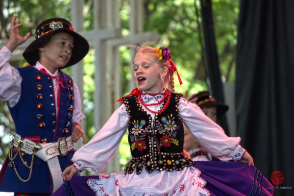 Medium full shot of two children dancing in colorful attire. The one on the left faces right with left hand on his waist and right-hand raised. He wears a white long-sleeve shirt, a long blue vest, a white belt, and a black hat. The girl at the image's center holds out her purple skirt. Her body faces the camera while her face looks slightly to the left. She wears a white long-sleeve shirt, a red necklace, and a vest embellished with beading and flower-shaped embroidery. Two other dancers are blurred in the background.