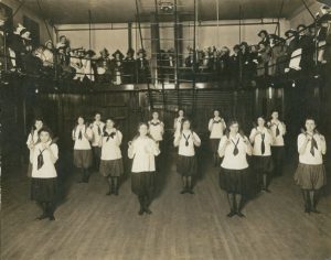 Sepia-colored long shot of fourteen young women in uniforms standing and facing the camera lens while demonstrating an exercise move. In the background, a group of people appear in an arched seating area on the upper floor. Some lean their body on the guardrails that enclosed the area. The room's floor, walls, and ceiling are visible.