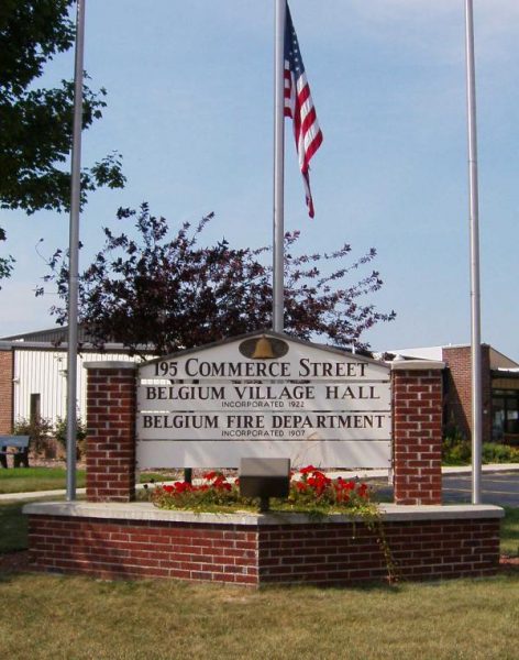 An outdoor monument sign mentions two facilities in the Village of Belgium: the Village Hall and the Fire Department. The monument sign also gives the address as 195 Commerce Street. The monument is built on brick walls decorated with red landscaping plants. Appearing in the middle behind the monument is a pole with an American flag.