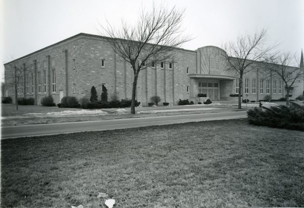 Wide shot of the long facade of Beth El Ner Tamid Synagogue in grayscale. The image shows two sides of the exterior wall. The left one features regularly spaced double rectangular windows. The right one has a covered entrance. Words in Hebrew are inscribed on top of the door and on its left and right sides.