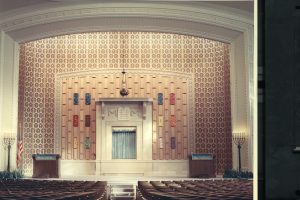 Interior of Congregation Emanu El B'ne Jeshurun. Rows of pews are visible in the foreground, with an aisle in the middle. A bimah with an ornate wall is in the background. A Hanukkah menorah stands on the platform's far left next to an American flag. Another menorah stands on the far right.