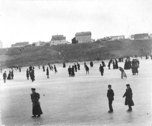 High-angle wide shot of people in winter clothes skating on the frozen Milwaukee River. Three young people are in the foreground, and many are scattered in the background. Several buildings appear atop the bluff in the distance.