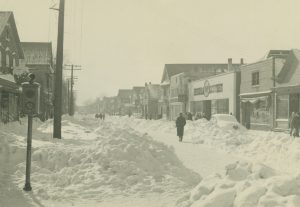 Sepia-colored long shot of Teutonia Avenue covered with snow. The road's middle part is plowed. Some people walk on it. Snowdrifts are piled up on most parts of the street, the sidewalk and some buildings' front areas on the left. A car partially buried in snow is parked on the street side on the right. Groups of people walk on the other sidewalk next to commercial buildings on the right.