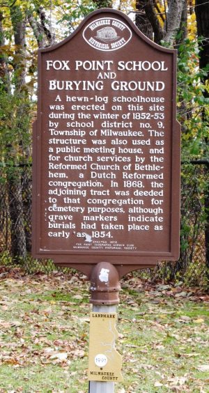 A standing historical marker of Fox Point School and Burying Ground in a brown-colored material contains the description of the building written in white paint. The sign is erected over a green lawn filled with scattered brown leaves. Below the sign is a metal plate in the shape of Milwaukee County and highlighting local rivers reading "Landmark 1991 Mmilwaukee County." Some feet behind are trees growing outside a chain link fence. The text of the marker reads "Fox Point School and Burying Ground. A hewn-log schoolhouse was erected on this site during the winter of 1852-53 by school district no. 9, Township of Milwaukee. The structure was also used as a public meeting house, and for church services by the Reformed Church of Bethlehem, a Dutch congregation. In 1868, the adjoining tract was deeded to that congregation for cemetery purposes, although grave markers indicate burials had taken place as early as 1854."