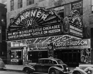 Exterior view of Warner Theater displaying a large marquee over its entrance. The marquee features signage of the theater, two movie titles and their stars' names, embellished by a series of glowing light bulbs. The lights on the marquee's underside illuminate two entrances beneath. The left entrance promotes "My Little Chickadee" movie with two big caricatures. The right one features "Castle on the Hudson" movie title and the casts' photos. Cars line the street side next to the theater.